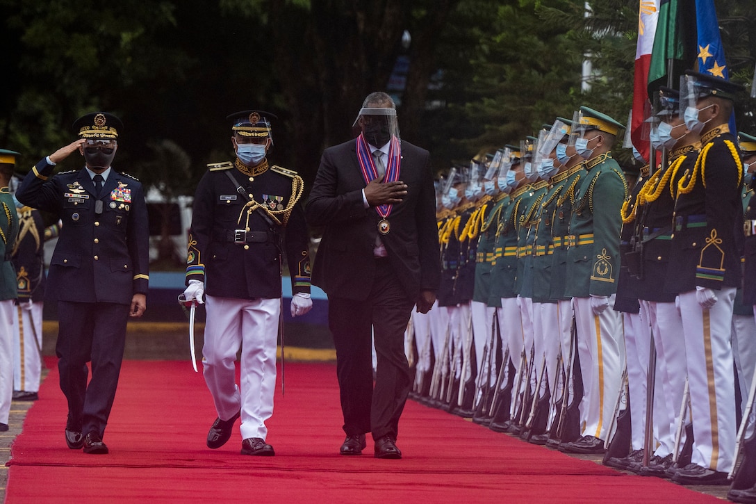 Several men walk along a red carpet in front of a row of service members.