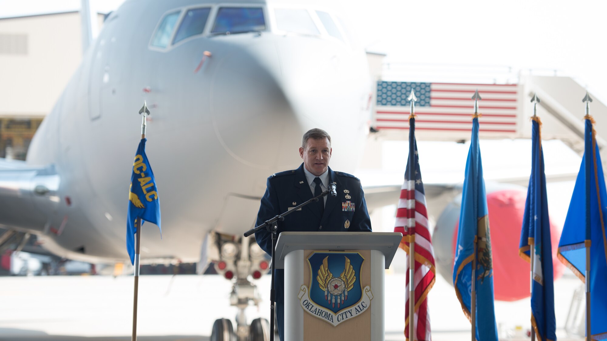 Man at podium with airplane and flags behind him.