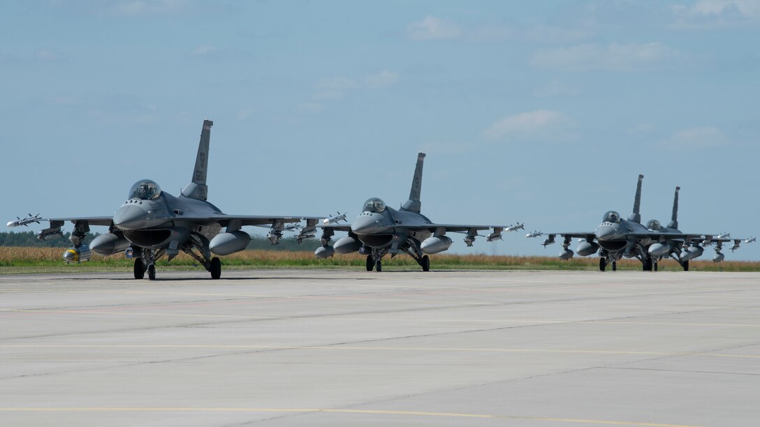Multiple military aircraft taxi down a runway in a row on a clear day