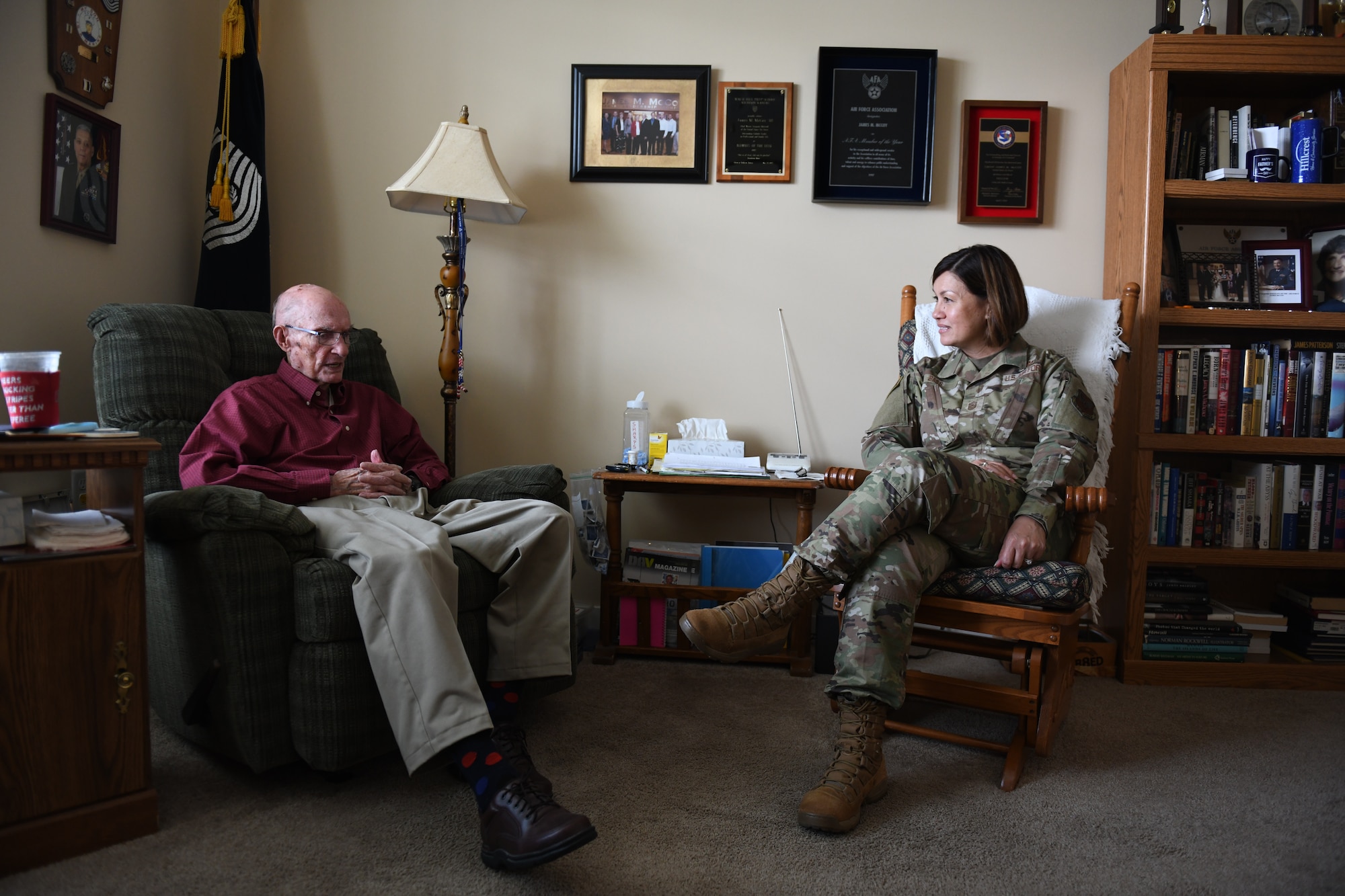 Retired Chief Master Sergeant of the Air Force James McCoy sits on the left in a recliner, and Chief Master Master Sergeant of the Air Force JoAnne S. Bass sits on the right in a wooden rocking chair facing each other in an office filled with awards on the wall, a bookshelf, a lamp and a flag.