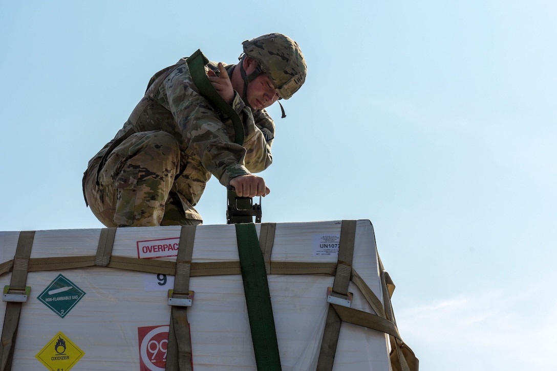 A soldier kneels atop a pallet and manipulates an attached strap.