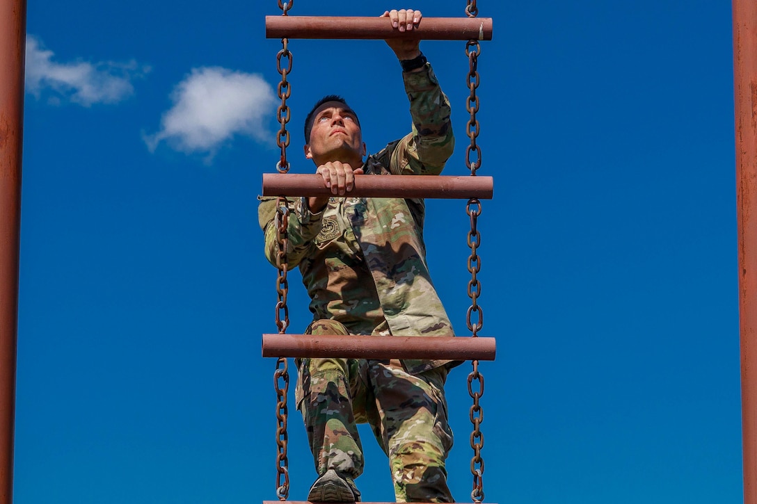 A soldier climbs an outdoor chain ladder, with blue sky as backdrop.