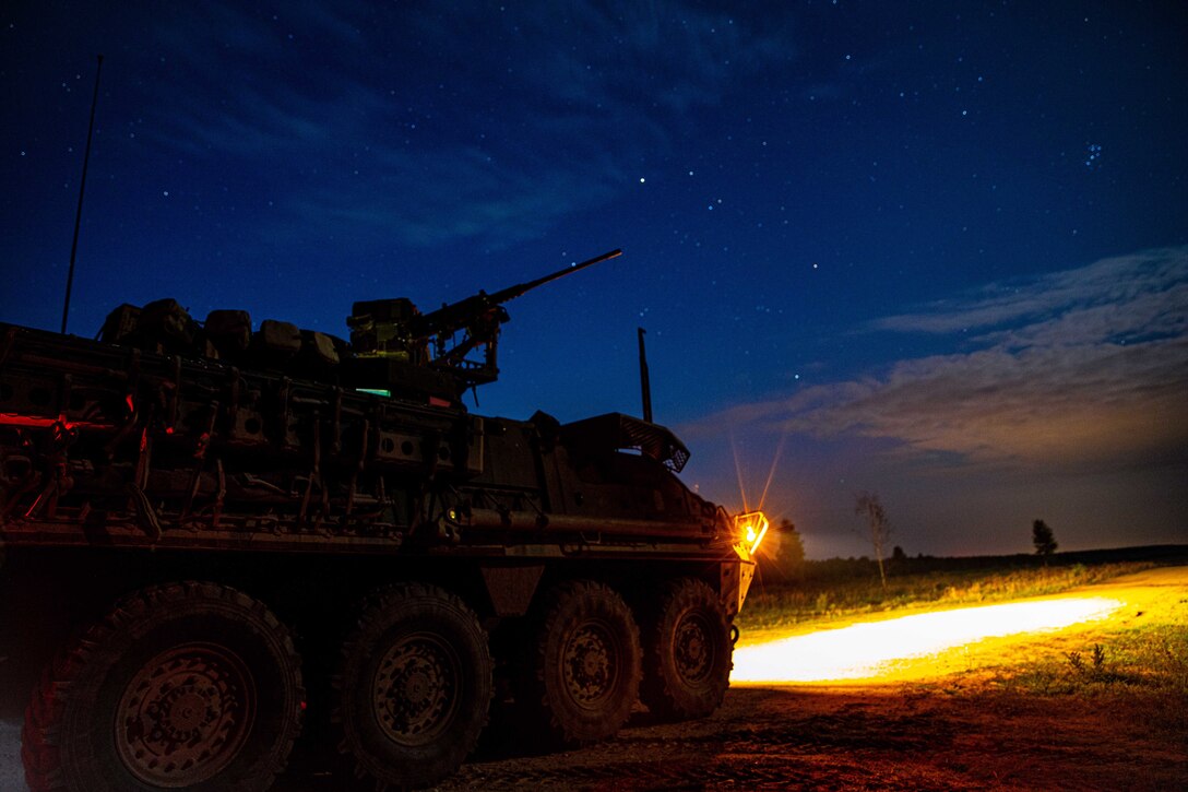 An Army vehicle with its headlights on sits in a field under a starlit sky.
