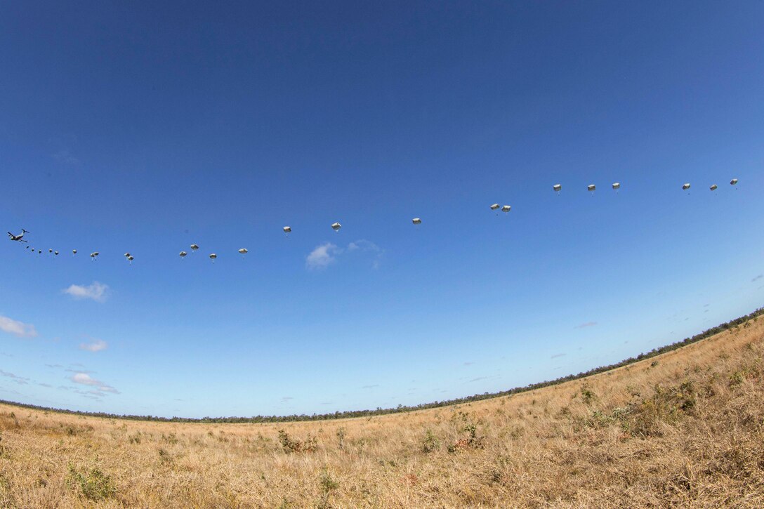 Soldiers jump out of an aircraft with parachutes over a field.
