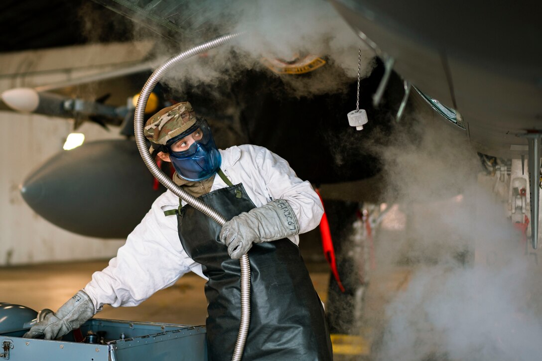 An airmen in protective gear holds a hose attached to an aircraft while smoke billows around.