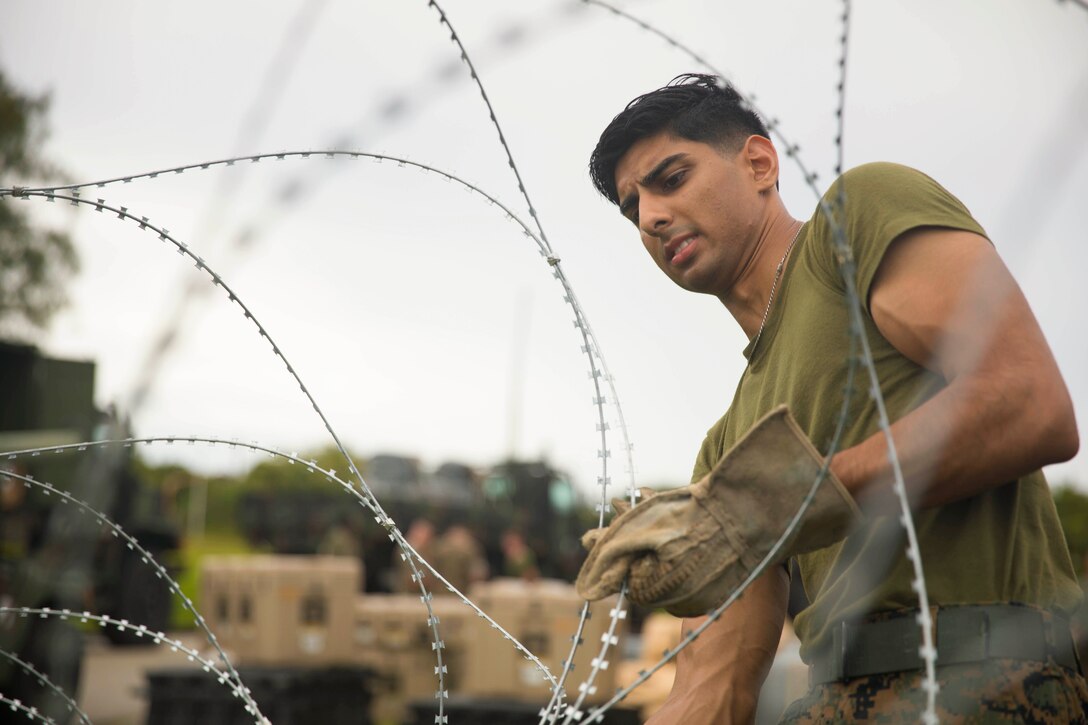 A Marine wearing gloves unravels barbed wire with military vehicles in the background.