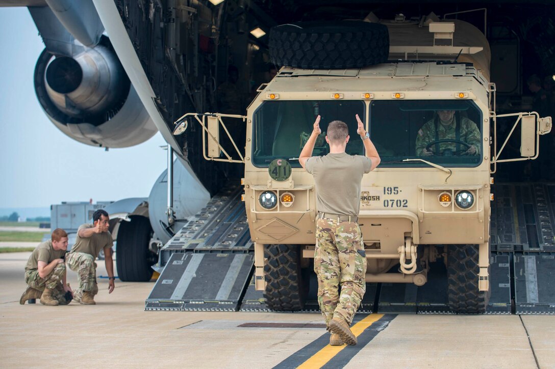 An airman guides another airman inside a vehicle into an aircraft as two others watch.
