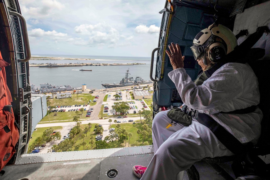 A woman waves from an open helicopter door at a moored ship below.