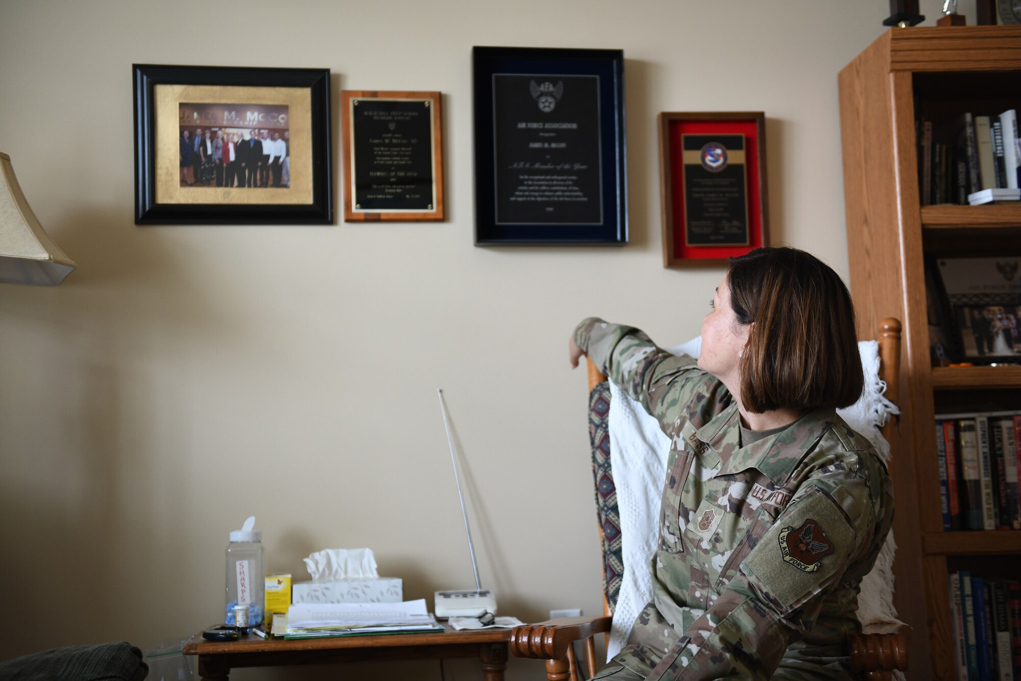 Chief Master Sergeant of the Air Force JoAnne S. Bass sits in a chair with her arm on the back rest turned over her right shoulder looking at the wall behind her. The wall behind her has four frames, one photo and three awards.