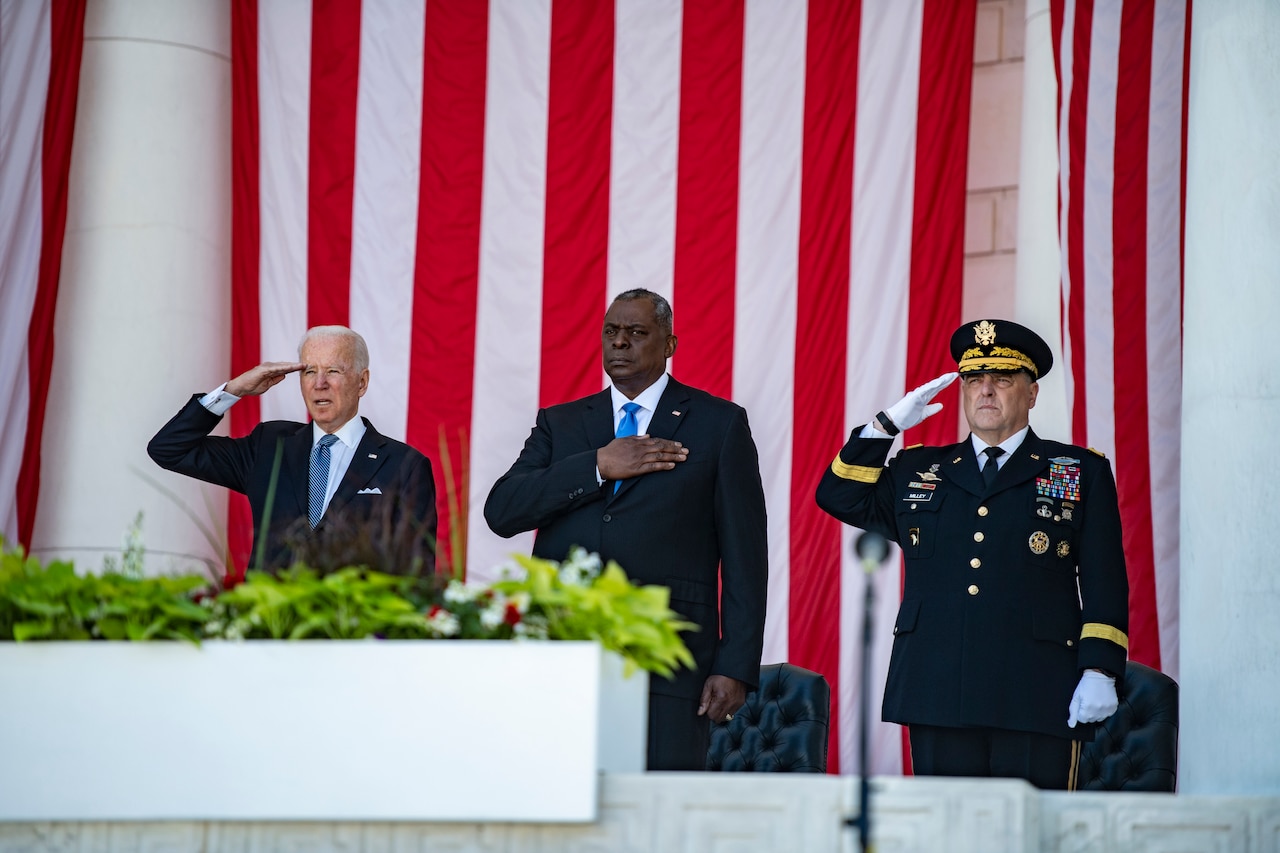 Three men stand at attention in front of large U.S. flags.
