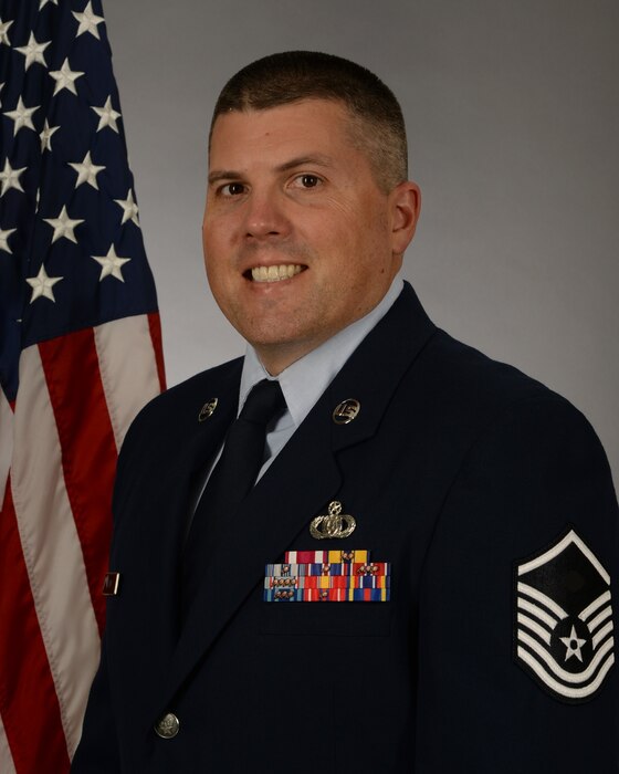 Official photo of MSgt Eric Frank, Superintendent of the Heritage of America Band, Langley AFB, VA.  MSgt Frank is wearing blue service dress in front of a grey background and the American flag.