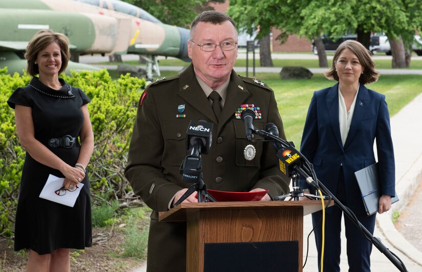 A man in military uniform speaks at a podium, two women in civilian clothes stand behind him.