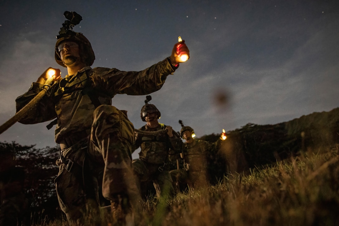 Soldiers walk through grass holding a light in one hand and a rope in the other.