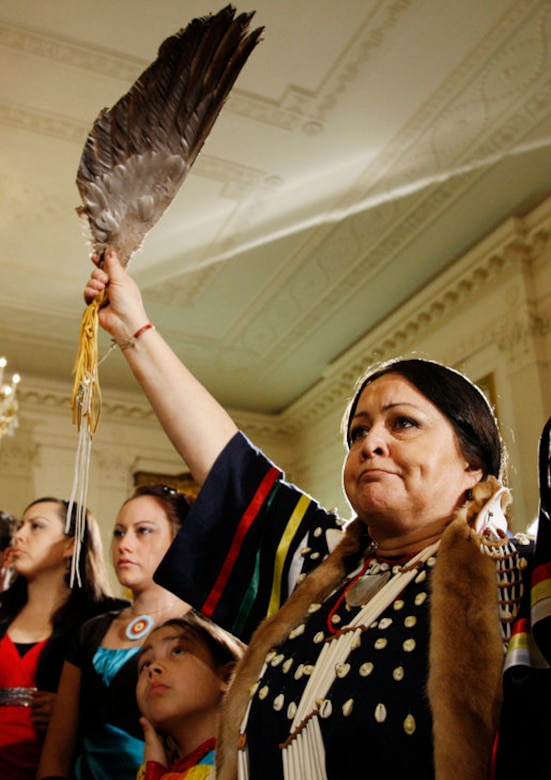 A woman dressed in Native American attire holds a large feather above her head.