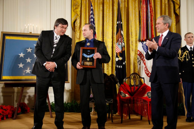 During a formal event, one man applauds as another man holds a shadowbox with a medal in it; a third man leans over to speak to the man holding the shadowbox.