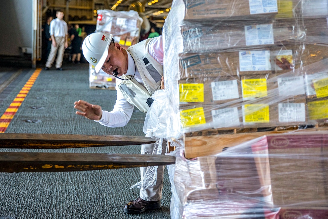 A sailor raises his hand and holds a whistle in his mouth while watching a forklift approach a pallet of cargo on a ship's deck.