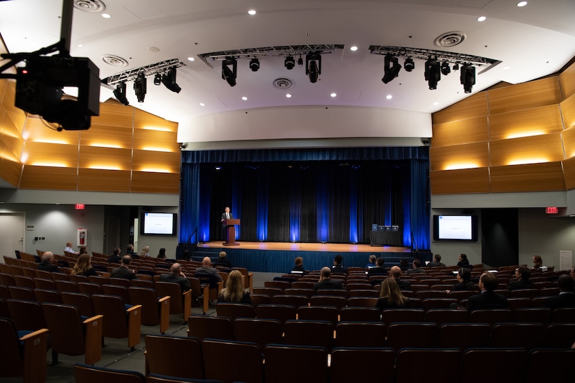 A man on a stage speaks to socially distanced people seated in an auditorium.