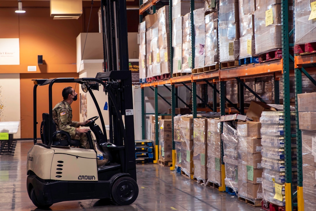 A soldier wearing a face mask operates a forklift to stack pallets of donated food and supplies