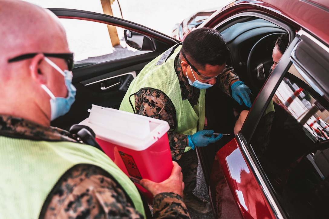A soldier wearing a face mask and gloves gives a person seated in a car a vaccine while another soldier wearing a face mask holds a hazard container for the syringes.