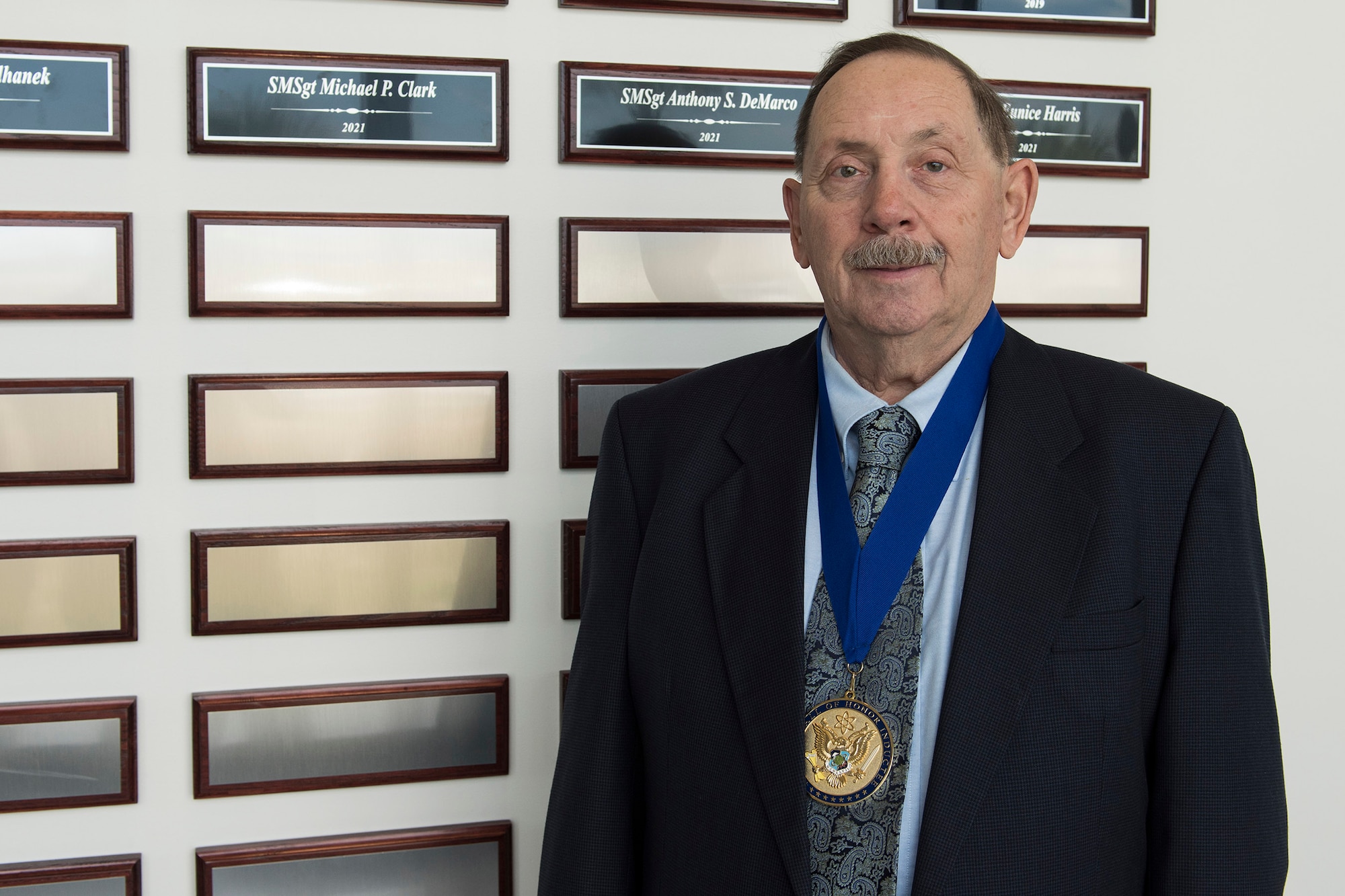 Tony DeMarco, a retired Air Force senior master sergeant, stands in front of his plaque after his induction to the Air Force Technical Applications Center's Wall of Honor during a ceremony May 26, 2021.  Seen around DeMarco's neck is the medallion that is presented to each honoree who, through the course of their career at AFTAC, demonstrated great character and whose actions truly discriminated them from thousands of other center employees, both military and civilian. (U.S. Air Force photo by Matthew S. Jurgens)
