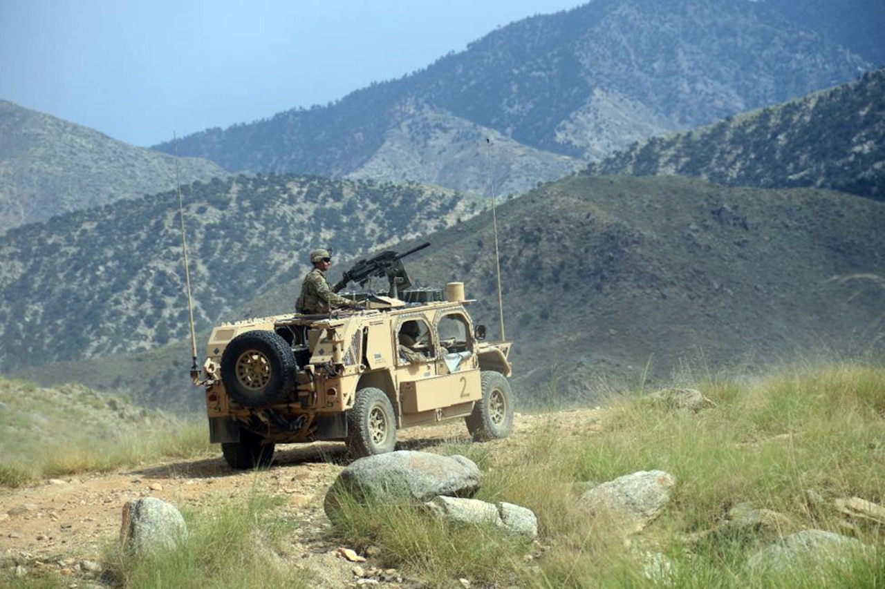 A combat vehicle along a gravel road with mountains in the background.  A soldier sits atop the vehicle near a gun that is mounted on the roof.