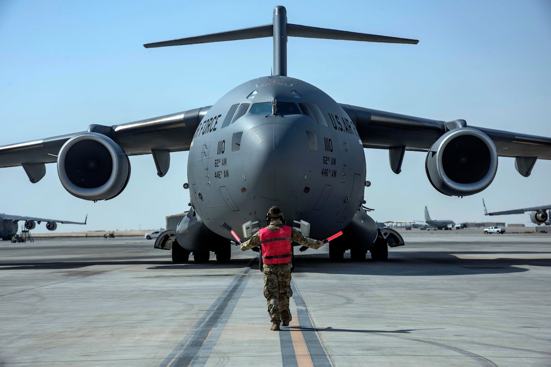 A large military aircraft is on an airfield. A service member uses hand wands to guide the aircraft.