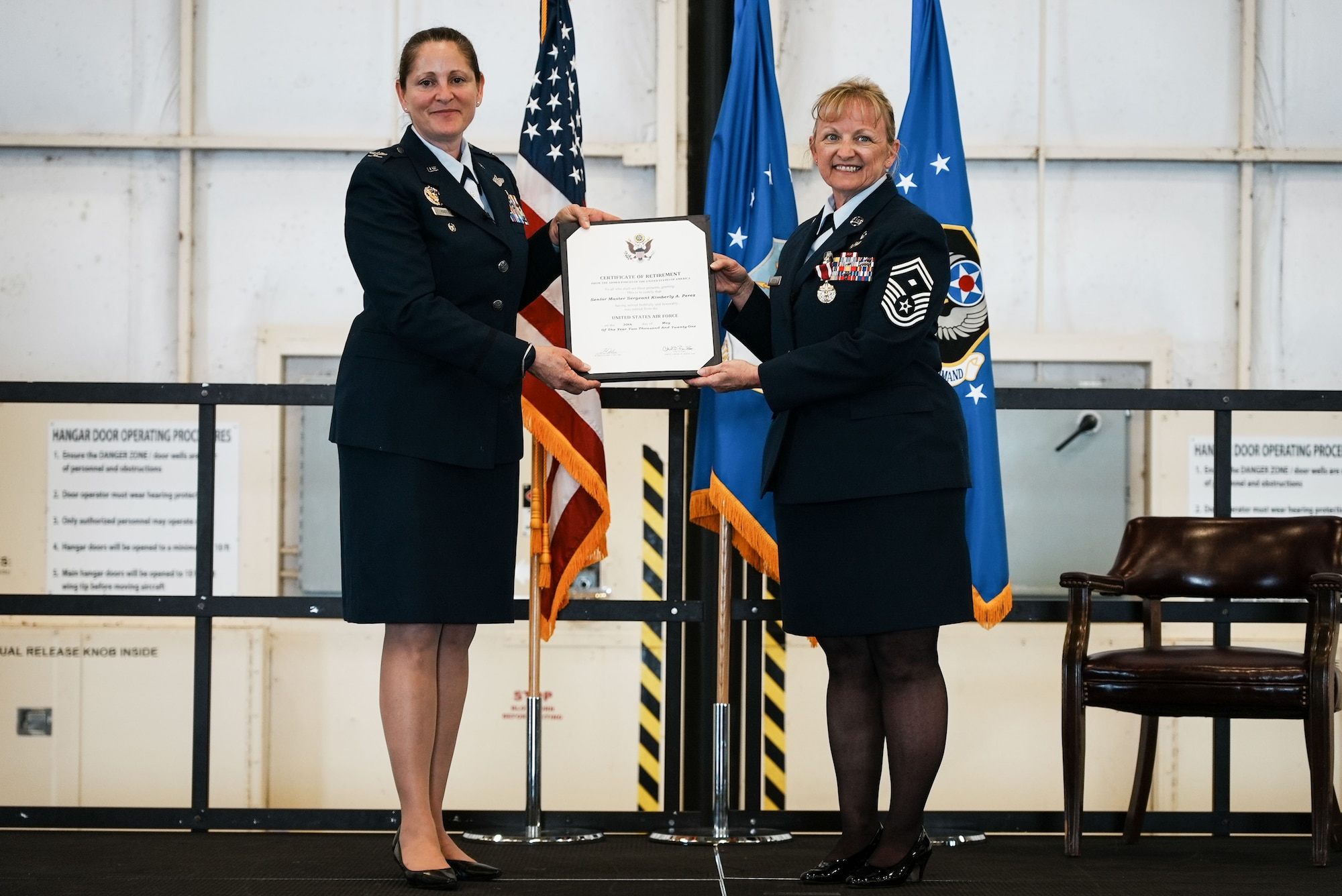 Two women in dress blues hold a certificate on a stage.