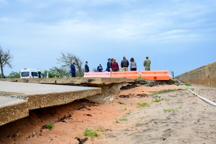 A photo shows the backs of eight people who are looking at something in the distance as another person appears to take notes. In the foreground, barricades block off an area where sand has eroded beneath a concrete slab.