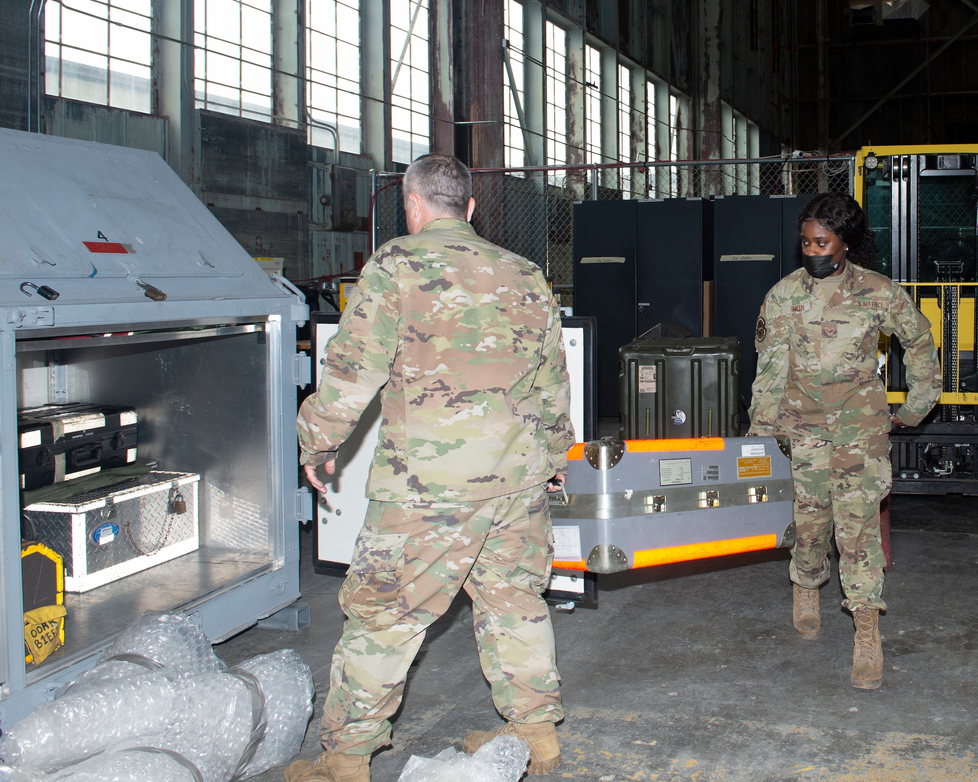 two Airmen carry a medium sized container; that contains test set to be packed into large metal kit; you can see inside metal kit equipment cases previously packed and bubble wrapped equipment on the floor ready for packing inside