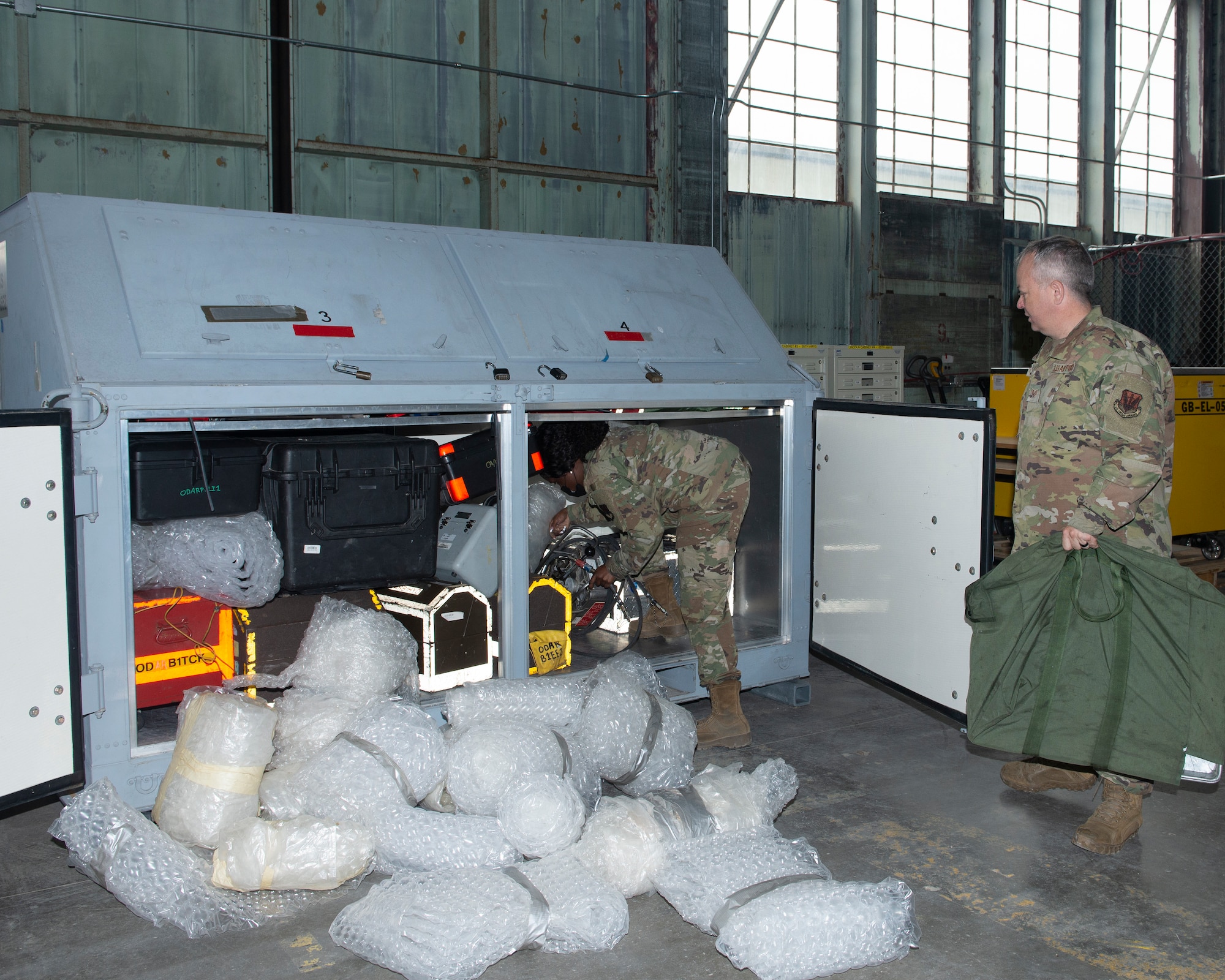 large metal box; with doors open; equipment being packed inside; bubble wrapped equipment in the foreground of photo; female Airman half inside large metal box situating a piece of equipment as male Airman waits to load equipment in duffle bag