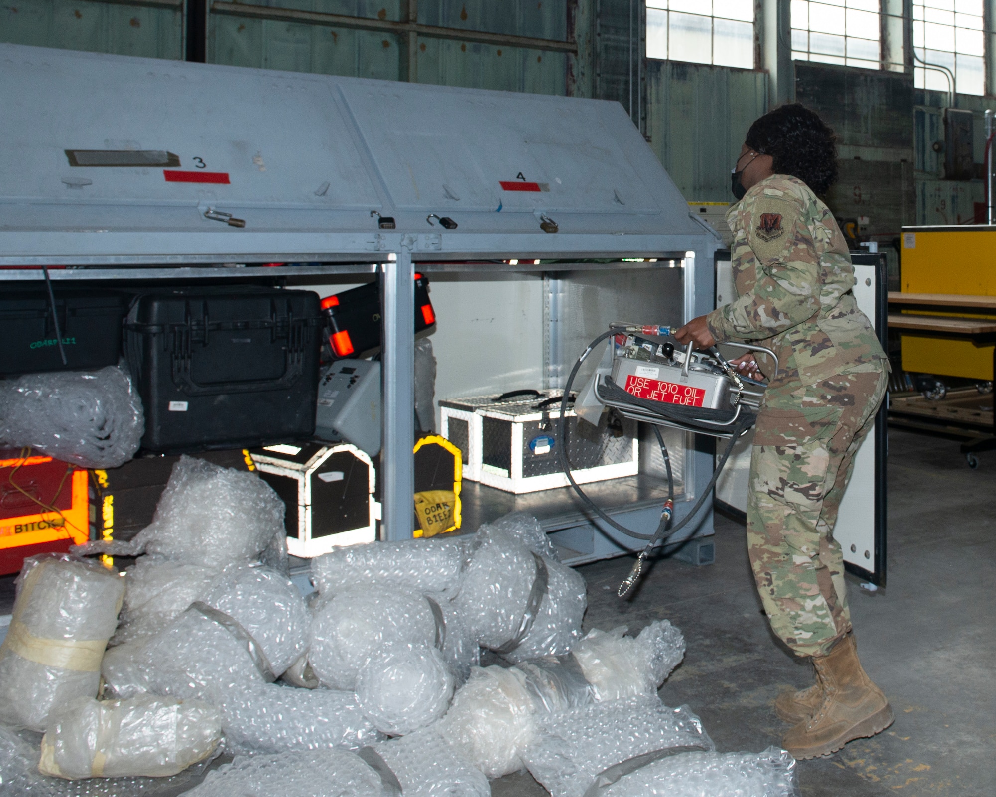 A female Airman carries a small metal box labled use 1010 oil or jet fuel. She is loading onto a large metal box which contains other equipment already packed. in the forefront of image are bubble wrapped equipment on the floor; waiting to also be loaded onto container