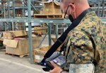 Guy in green camo uniform stands in a warehouse with shelves and boxes wearing a camera around his neck.