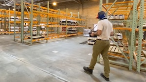 Man walks through a warehouse with boxes and shelves wearing a camera around his neck.