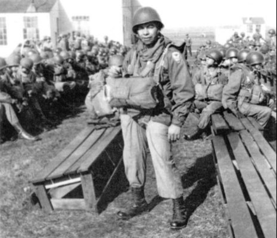 A man wearing a front-loaded parachute stands in the foreground with other men seated on benches behind him.