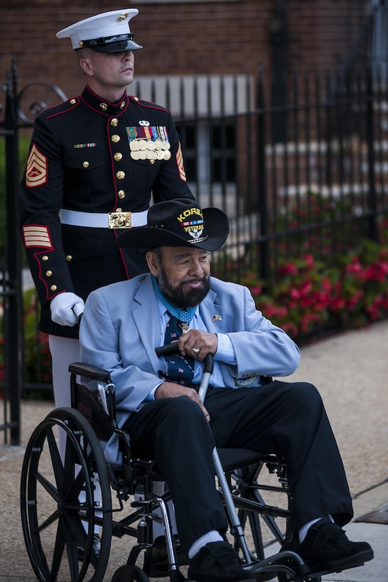 A Marine in dress uniform pushes the wheelchair of an older man in a cowboy hat.
