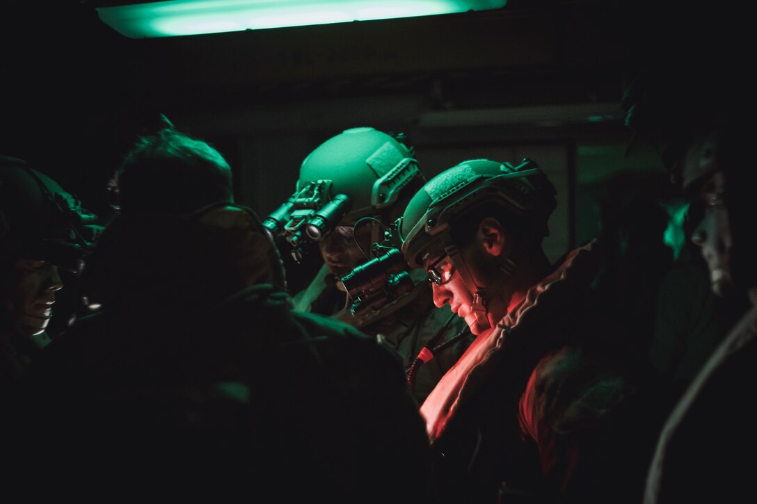 Marines wearing raid gear gather in a dark room illuminated by a small red light.