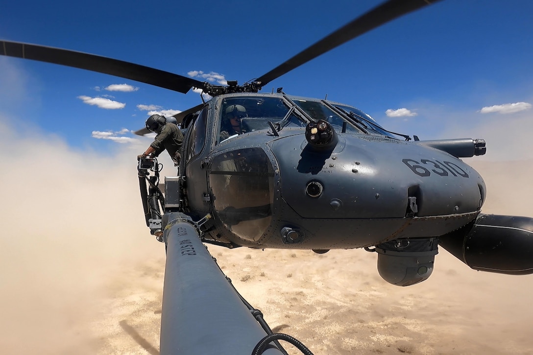 A service member leans out of an open helicopter that is hovering over desert terrain in a blue sky.