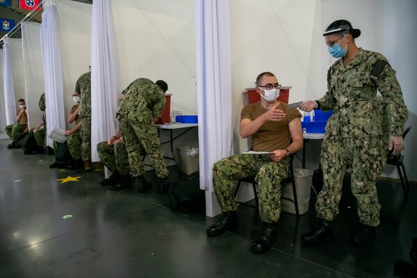 Inside a row of cubicles, a service member wearing a face mask and gloves hands a paper to a young service member who is seated and wearing a face mask. Similar activities are going on inside four other cubicles.