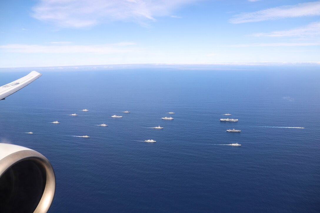 About 17 U.S. and foreign Navy ships travel in formation in blue seas, as part of an aircraft is visible flying nearby.