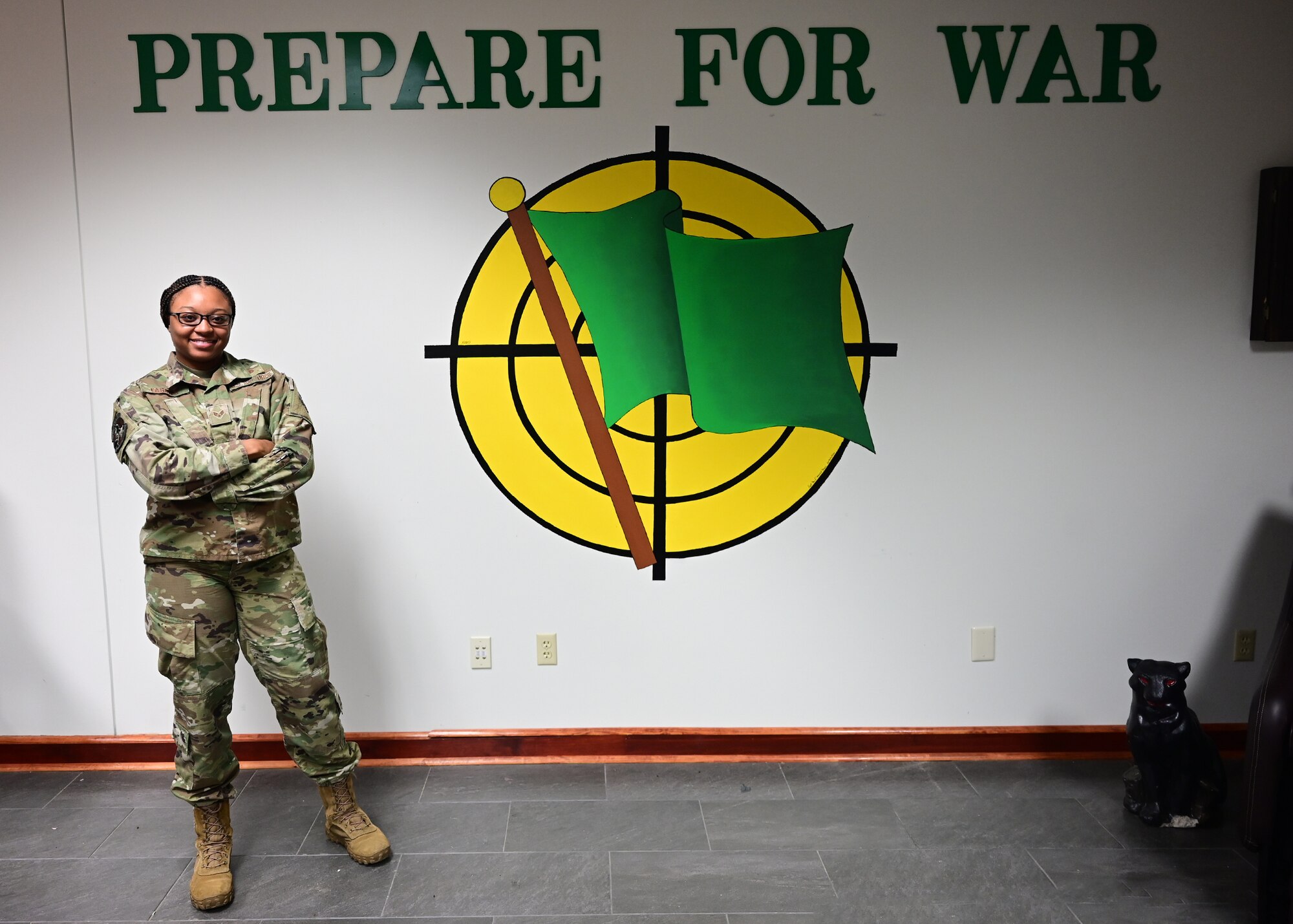 An airman stands in front of a wall with the text 'prepare for war' and a green flag logo.