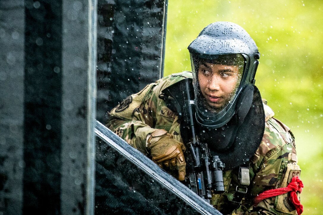 An airman wearing a protective helmet holds a weapon by a structure outside in the rain.