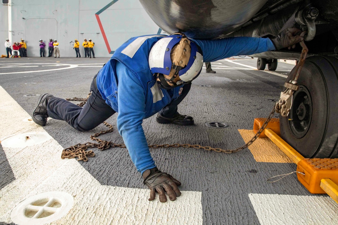 A sailor lays on the ground to secure chains to an aircraft.