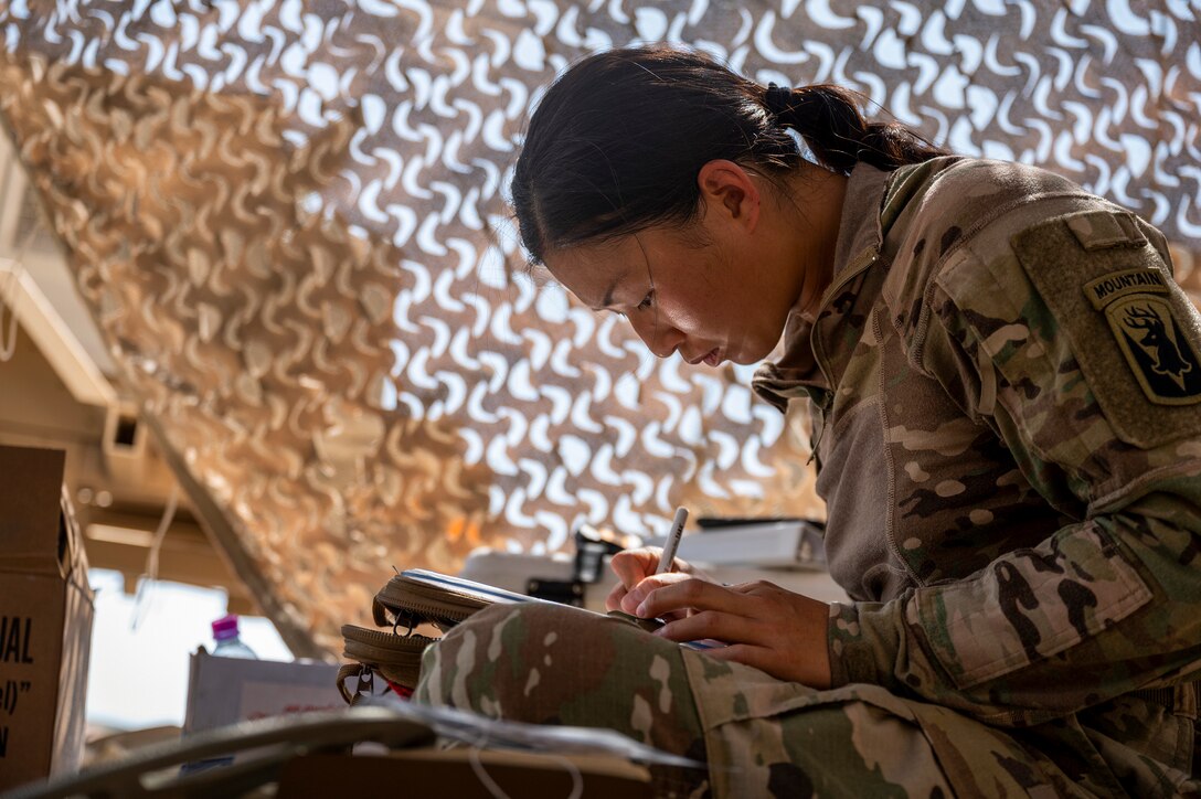 A soldier writes while sitting under protective netting.