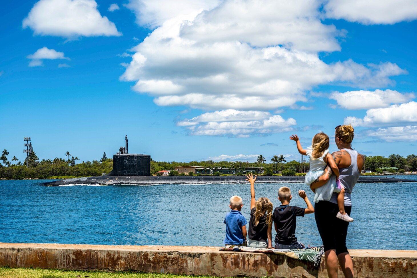 A submarine sails in blue waters as an adult and four young children watch and wave from the shore.
