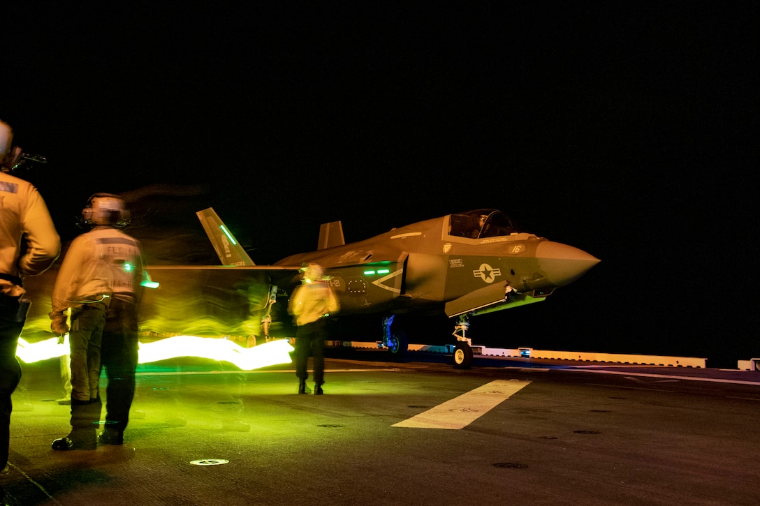 Sailors stand near an aircraft on a ship’s deck illuminated by green light.