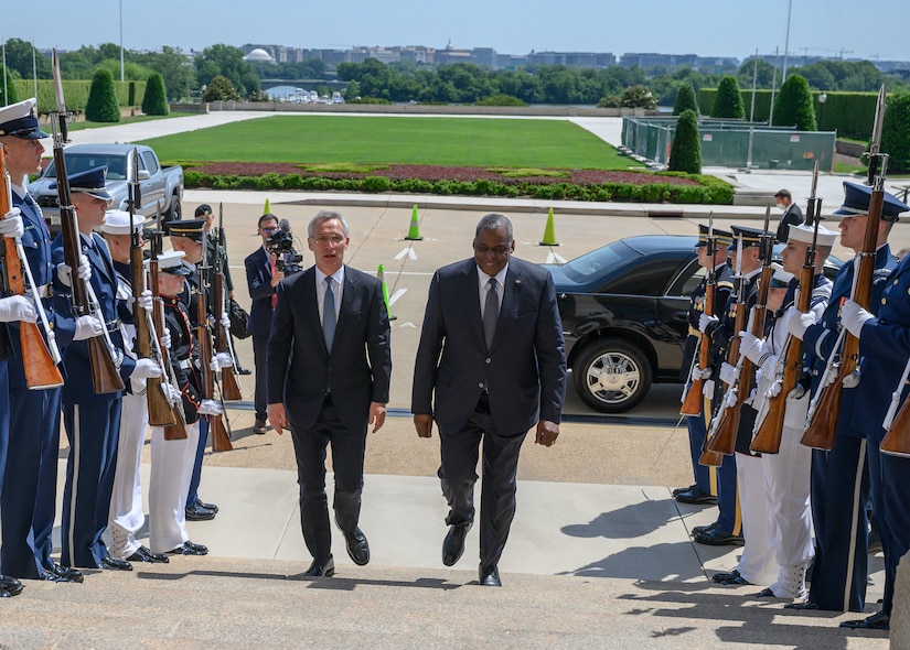 Two men in suits walk up stairs between rows of military service members.