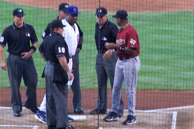 Six men stand on a baseball field and talk during a game; two men are wearing team uniforms.