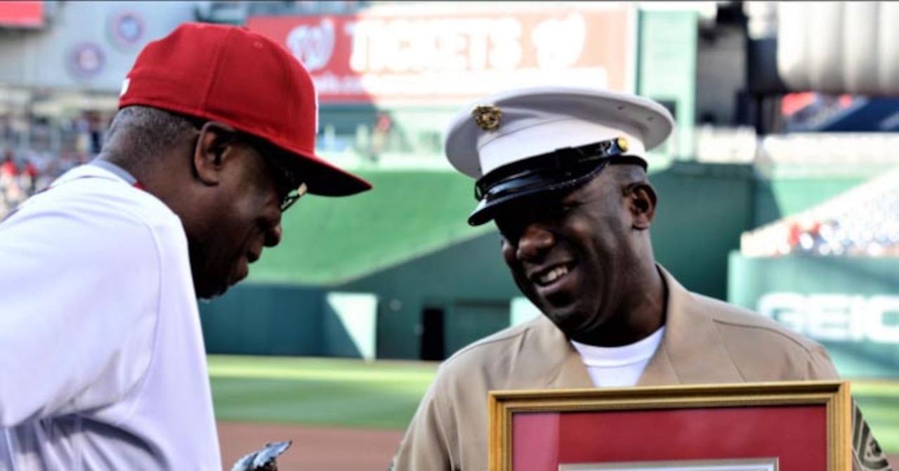 A man wearing a baseball uniform looks at a framed document being held by a man wearing a military uniform.