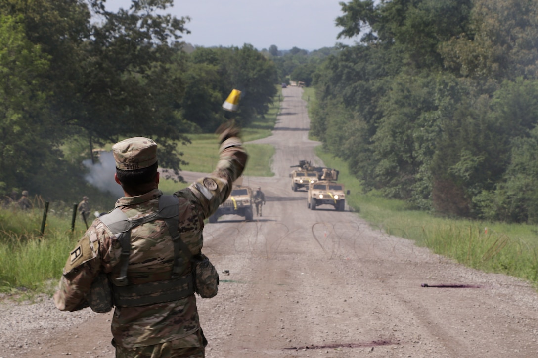 A soldier throws a smoke canister towards military vehicles on a dirt paved road in the woods.