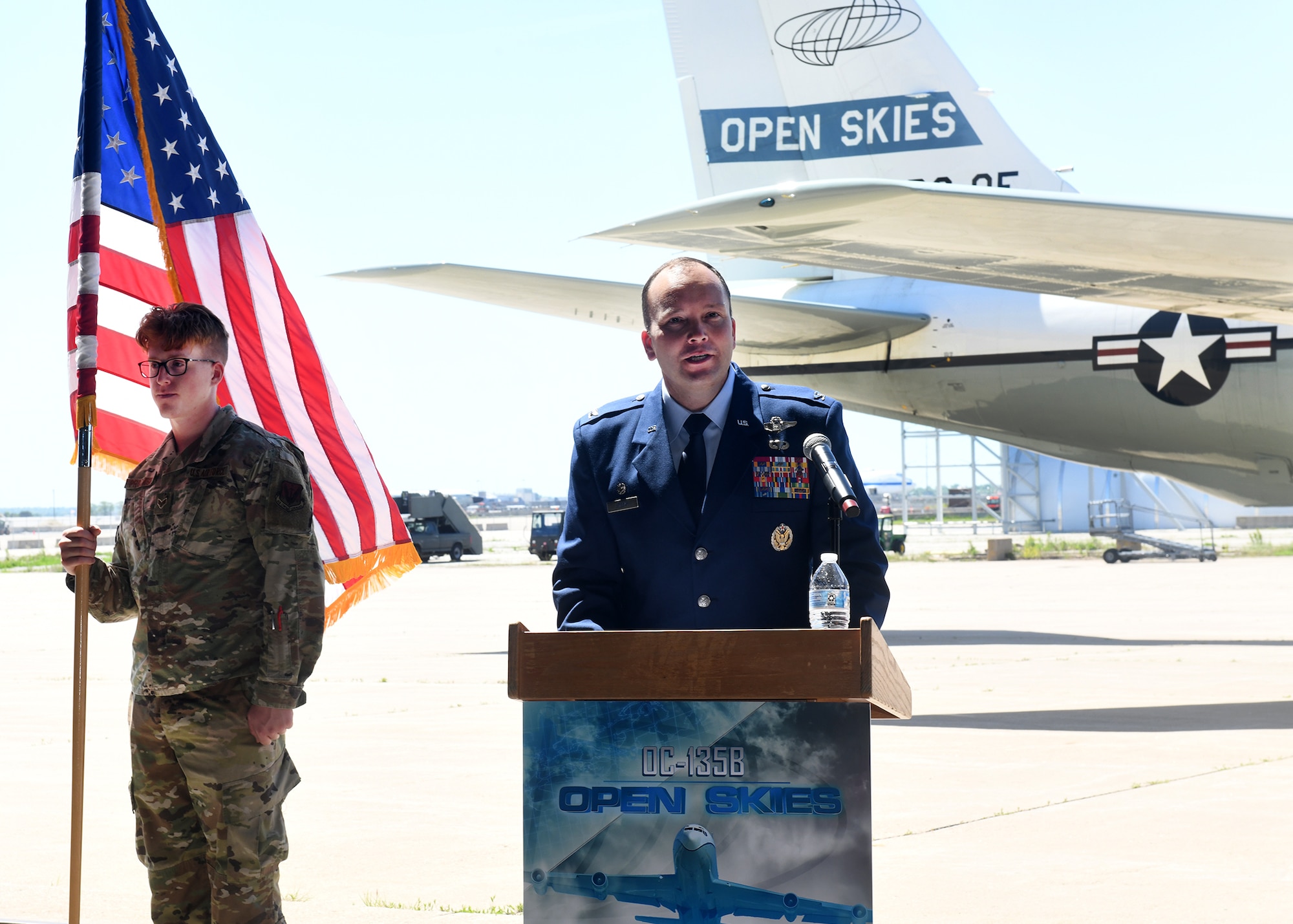 Man in Air Force uniform standing at a podium gives a speech. An Airman stands nearby holding the U.S. flag and the retiring aircraft is in the background.
